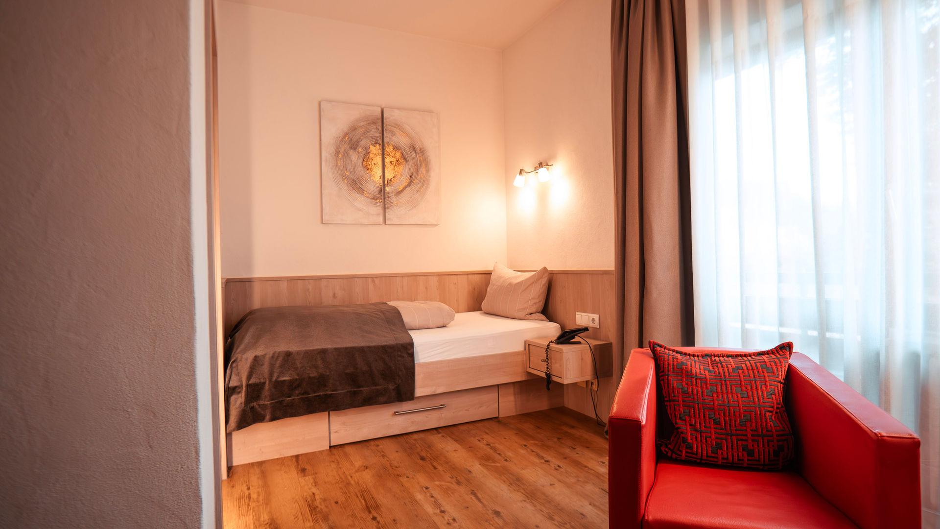 Gemütliches Einzelzimmer mit Bett, Holzfußboden, rotem Sessel und Vorhängen.