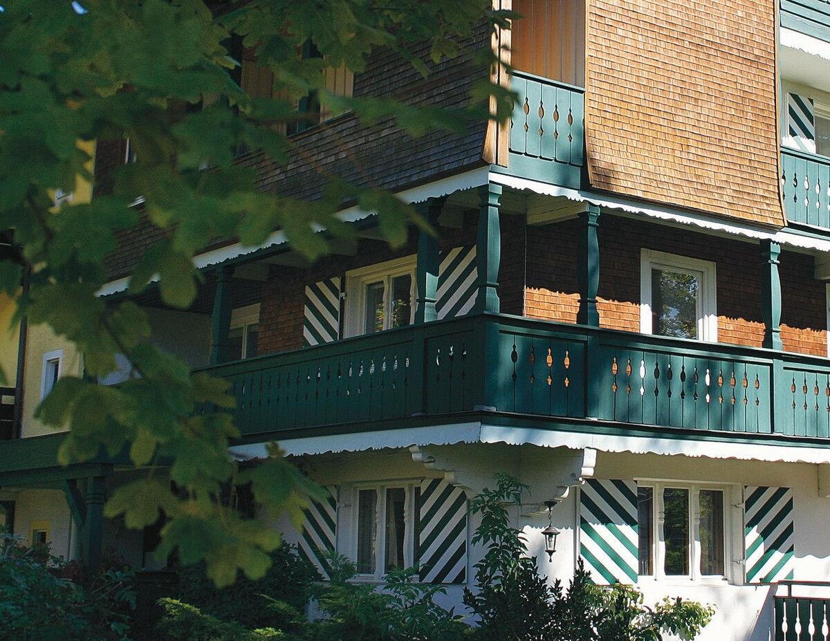 Hotelfassade mit grün gestrichenem Holzbalkon, umrahmt von Laub.