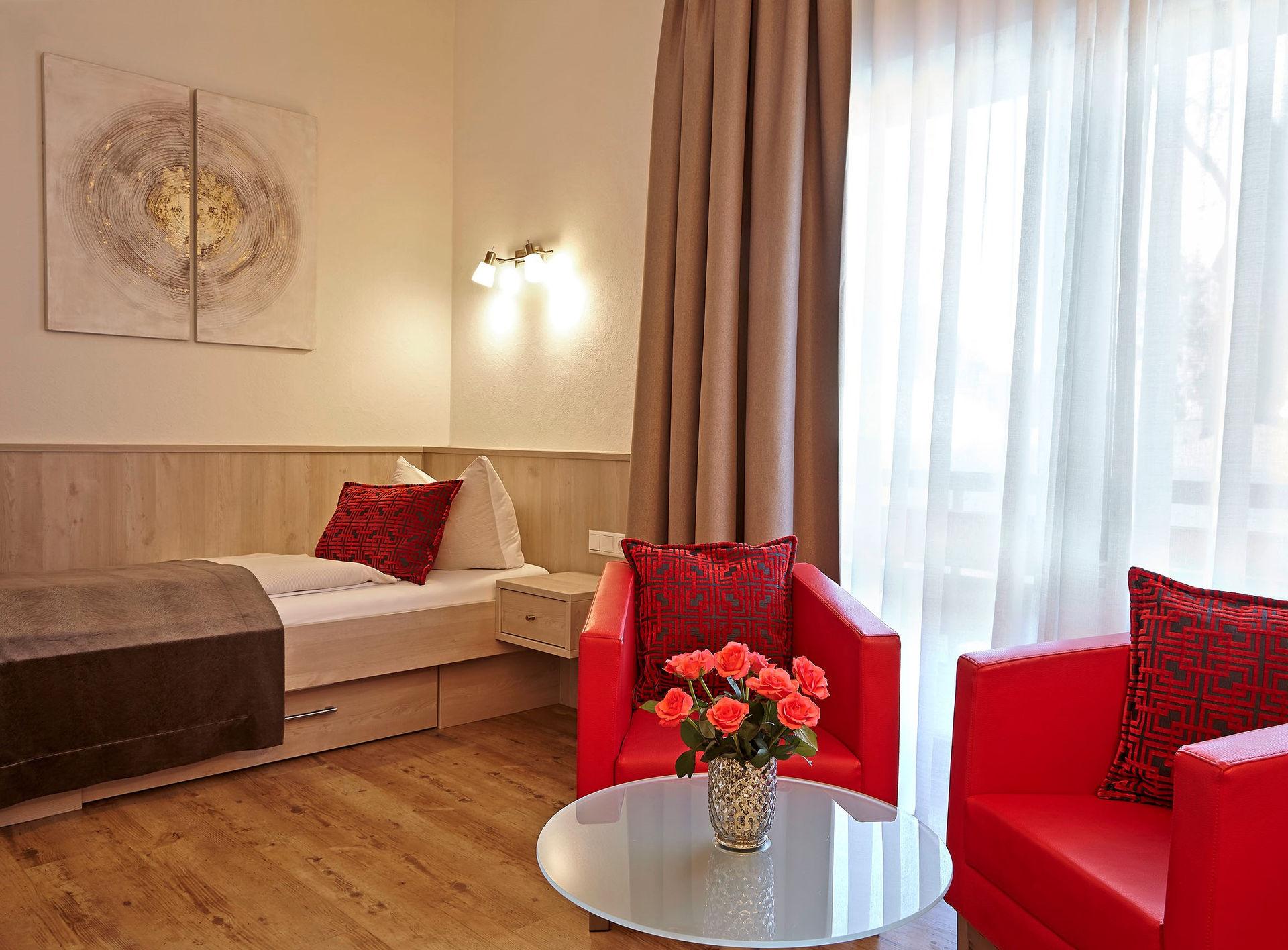 Gemütliches Hotelzimmer mit Einzelbett, roten Sesseln, Blumen und großem Fenster.