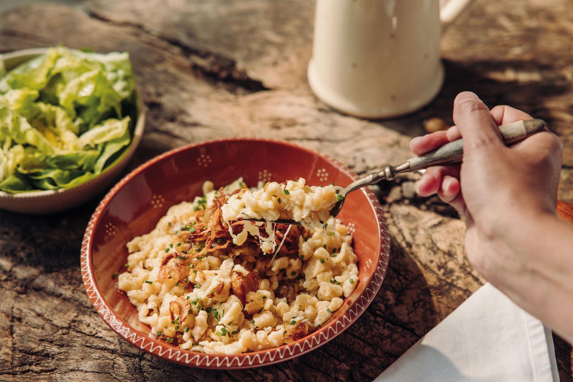 Hand isst herzhafte Käsespätzle mit Röstzwiebeln und frischem Salat.
